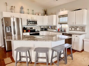 A kitchen with white cabinets, stainless steel appliances, a granite countertop, and three bar stools. - Experience oceanfront luxury at Pirate's View in Oak Island—perfect for 16 guests! (Oak Island)