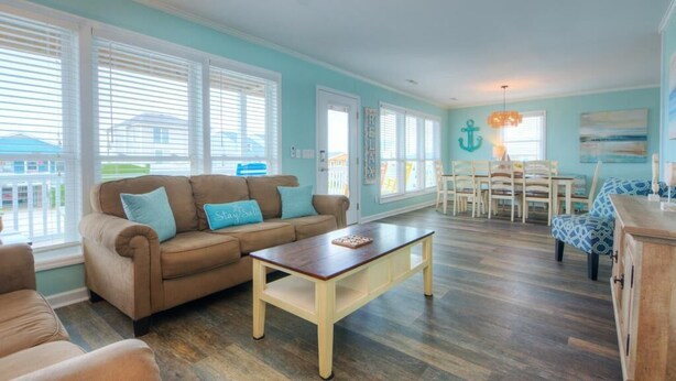 A living room with a brown sofa, a wooden coffee table, and a dining area with a chandelier. - Discover ocean views at Salt Shack, Oak Island! Perfect for families and pets, sleeps 8. (Oak Island)