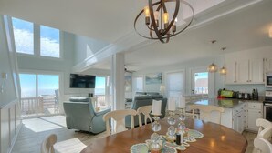 Ett rymligt vardagsrum med ett matbord för sex personer, ett stort fönster med utsikt över havet, en mysig sittgrupp med soffa och fåtöljer samt ett kök med vita skåp och en platt-TV.