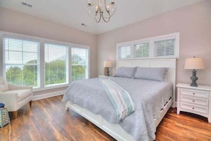 A bedroom with a bed, two bedside tables, a chair, and large windows with blinds.