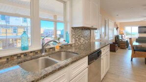 A kitchen with granite countertops, white cabinets, a tiled backsplash, and a large window overlooking a residential area. - Seas the Moment: 5BR oceanview retreat in Oak Island, perfect for families! (Oak Island)