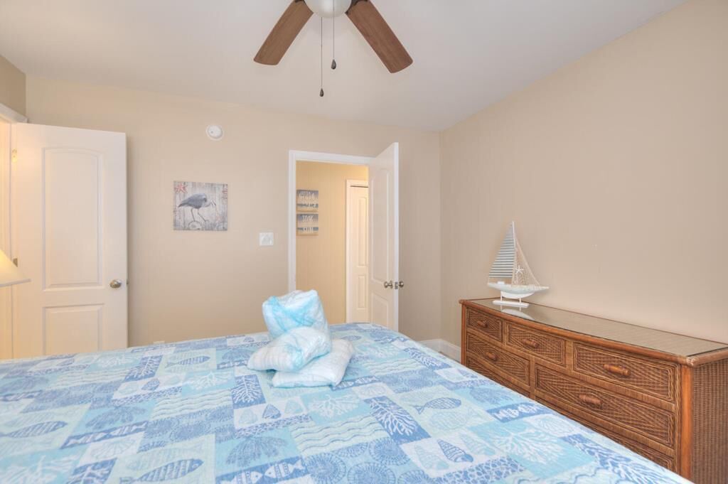 A bedroom with a bed, a wooden dresser, a ceiling fan, and a door leading to another room.