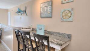 A dining area with a marble countertop, black chairs, and wall art including a 'Beach Life' sign and a shell decoration. - Seas the Moment: 5BR oceanview retreat in Oak Island, perfect for families! (Oak Island)