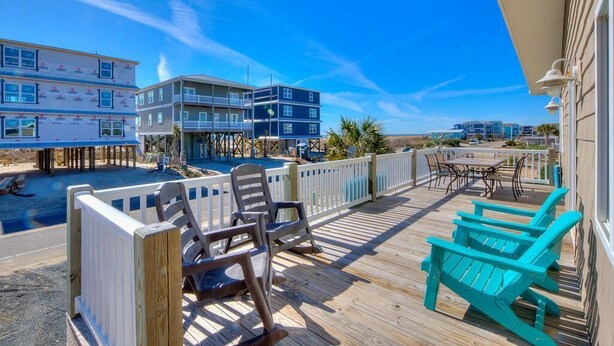 A wooden deck with chairs and a table, overlooking a beach and buildings. - Seas the Moment: 5BR oceanview retreat in Oak Island, perfect for families! (Oak Island)