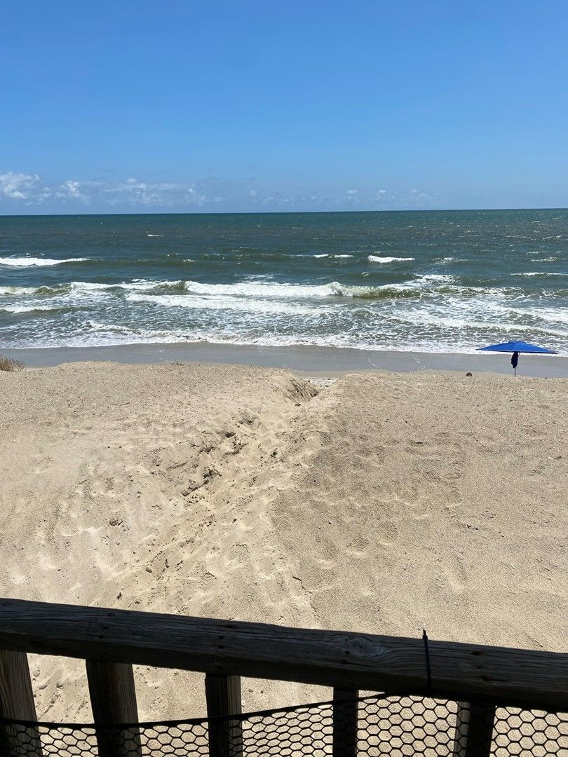 Une plage avec des vagues, une clôture en bois et une personne tenant un parasol bleu.