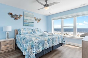 Un dormitorio con cama, mesita de noche, lámpara, ventilador de techo, ventana con vista al mar y decoración en la pared.