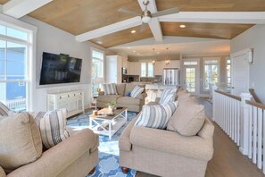 A spacious living room with a large TV, multiple seating areas, and a kitchen in the background. - Oceanview luxury awaits at Southern Tides, Oak Island – 5BR, pool, and family fun! (Oak Island)
