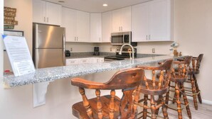 Une cuisine moderne avec des armoires blanches, des appareils en acier inoxydable et un comptoir en granit. On y trouve des tabourets de bar en bois et un panneau décoratif au mur.