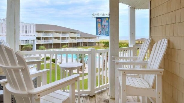 Un espace extérieur abrité, avec des chaises blanches et une petite table, donnant sur une piscine et un bâtiment doté de nombreux balcons.