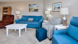 Un salon avec un canapé bleu, une table basse blanche, des fauteuils à rayures et un meuble en bois sur lequel est posé un panneau indiquant « PLAGE ».