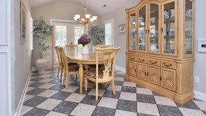Uma sala de jantar com chão xadrez, uma mesa de madeira com cadeiras, um lustre e um armário de vidro com louças.