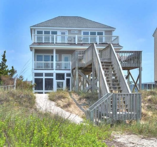 A two-story beachfront house with a wooden staircase leading to a balcony.
