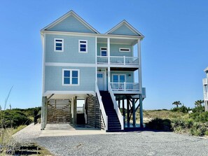 Une maison de deux étages située en bord de mer, dotée d’un balcon au deuxième étage, d’un escalier menant à l’entrée et d’un espace abrité à la base.