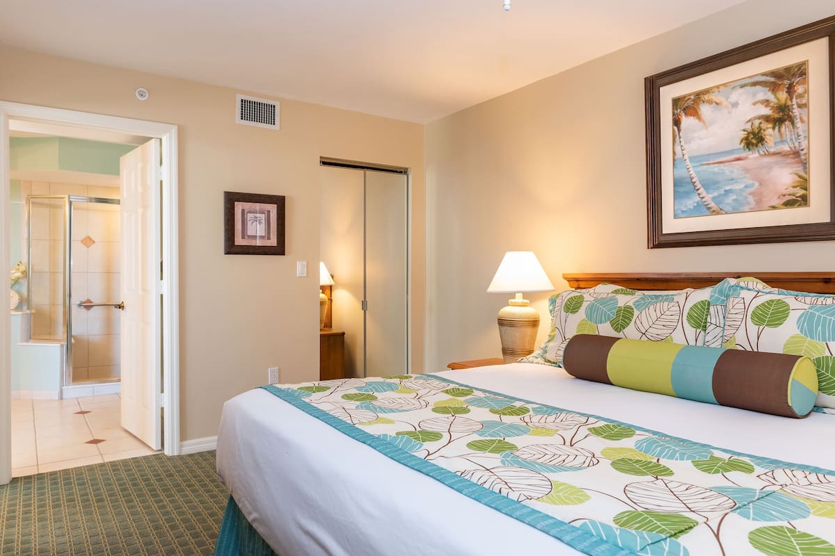 A hotel room with a bed, a nightstand, a lamp, a framed picture on the wall, and a door leading to a bathroom.