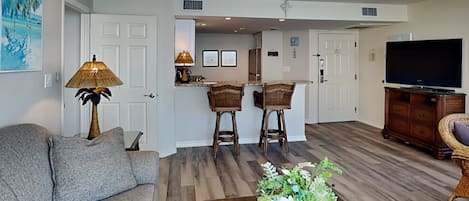 A living room with a sofa, a coffee table with a plant, a TV, and a kitchen area with bar stools.