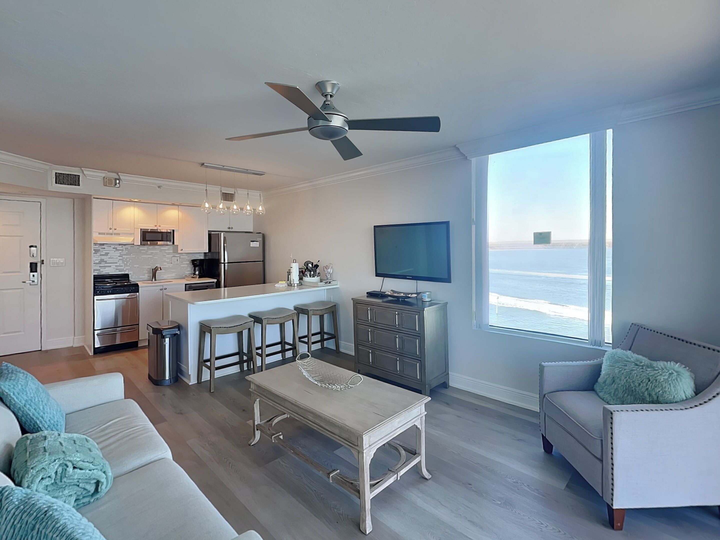 A modern kitchen with a central island, a dining area with a table and bar stools, a living room with a sofa and armchair, and a large window with a view of the ocean.