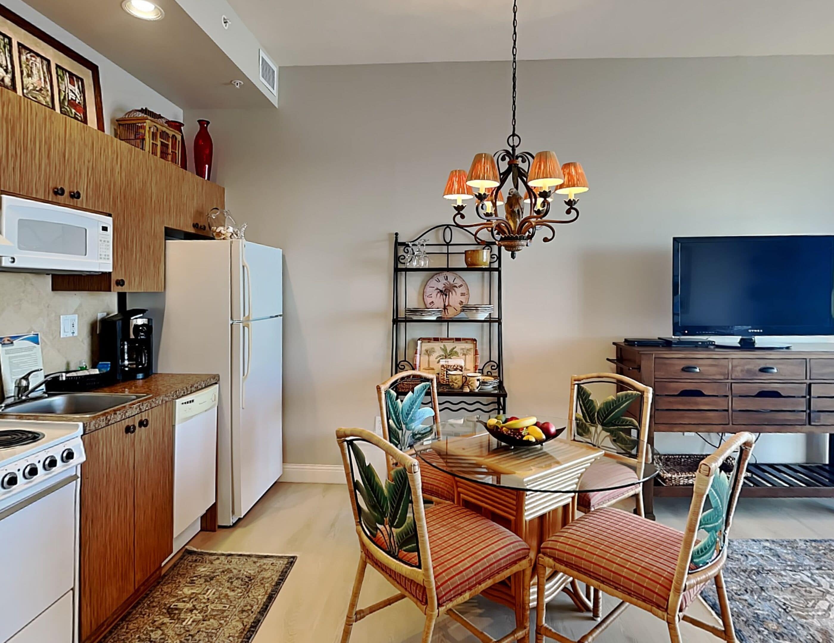 A modern kitchen with a dining area, featuring a central island, chairs, a chandelier, and a television.