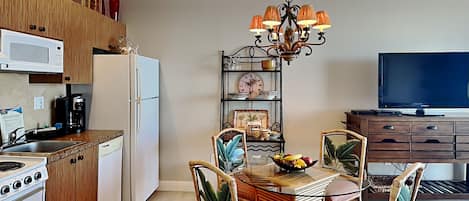 A modern kitchen with a dining area, featuring a central island, chairs, a chandelier, and a television.