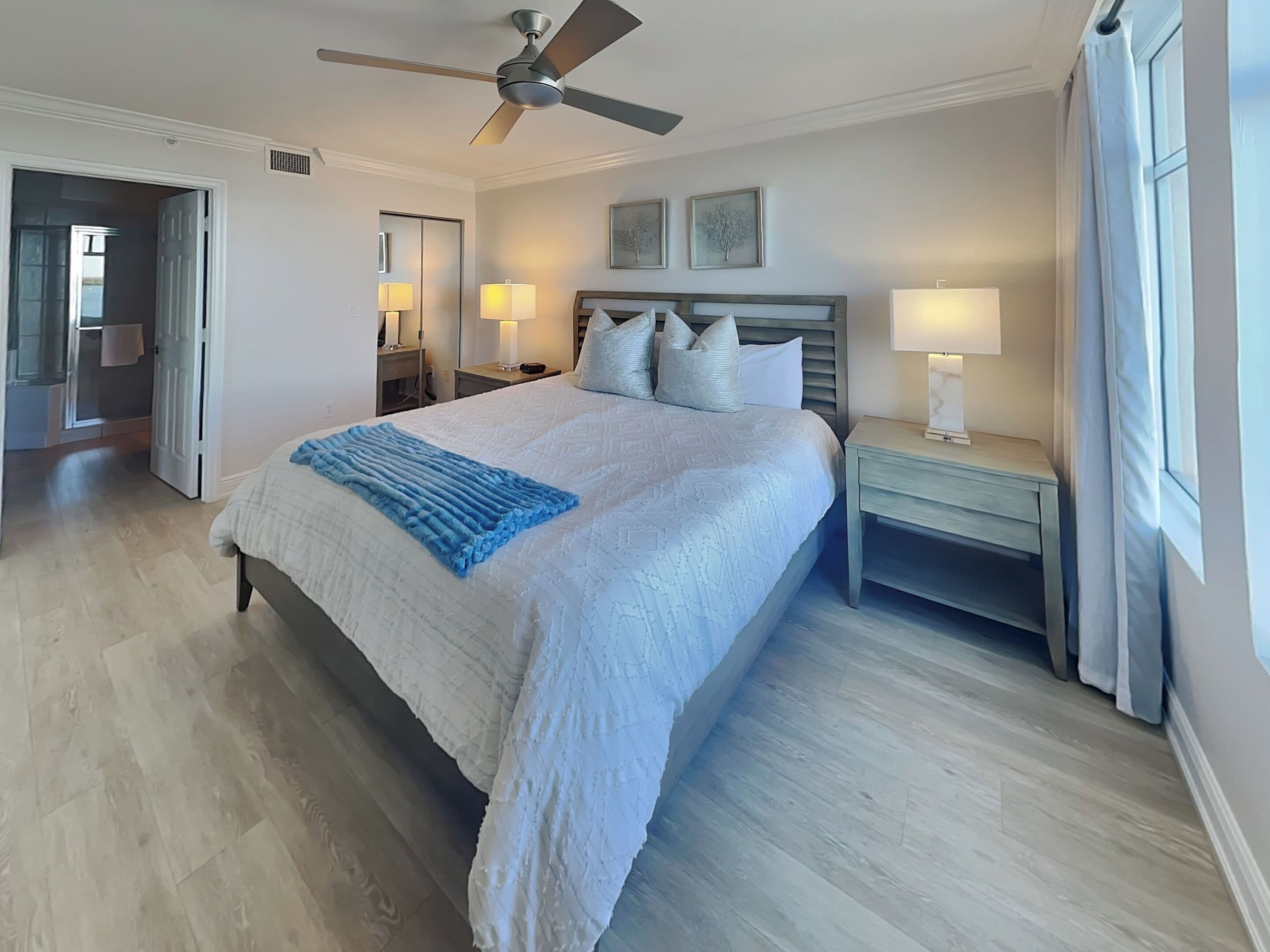 Un dormitorio con cama, mesita de noche, ventilador de techo y ventana con cortinas.