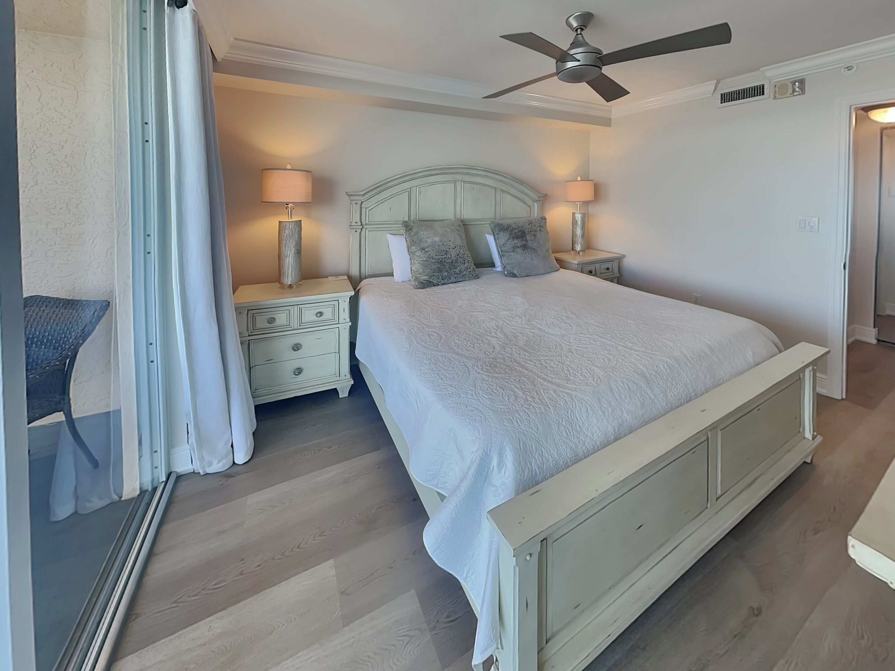 A bedroom with a bed, nightstands, lamps, a ceiling fan, and a view of the outside.