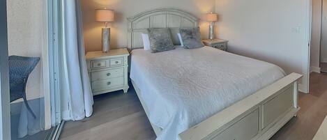 A bedroom with a bed, nightstands, lamps, a ceiling fan, and a view of the outside.