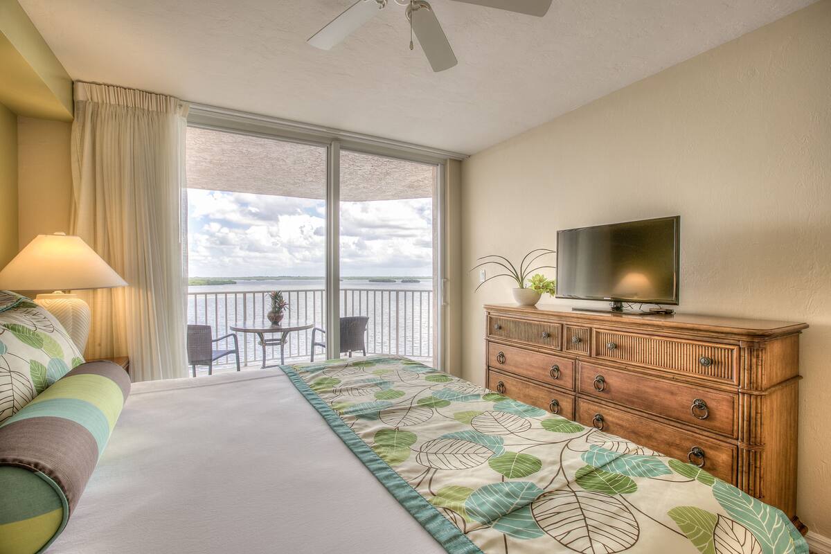 A hotel room with a bed, a television on a wooden dresser, a balcony with a view, and a table with chairs.