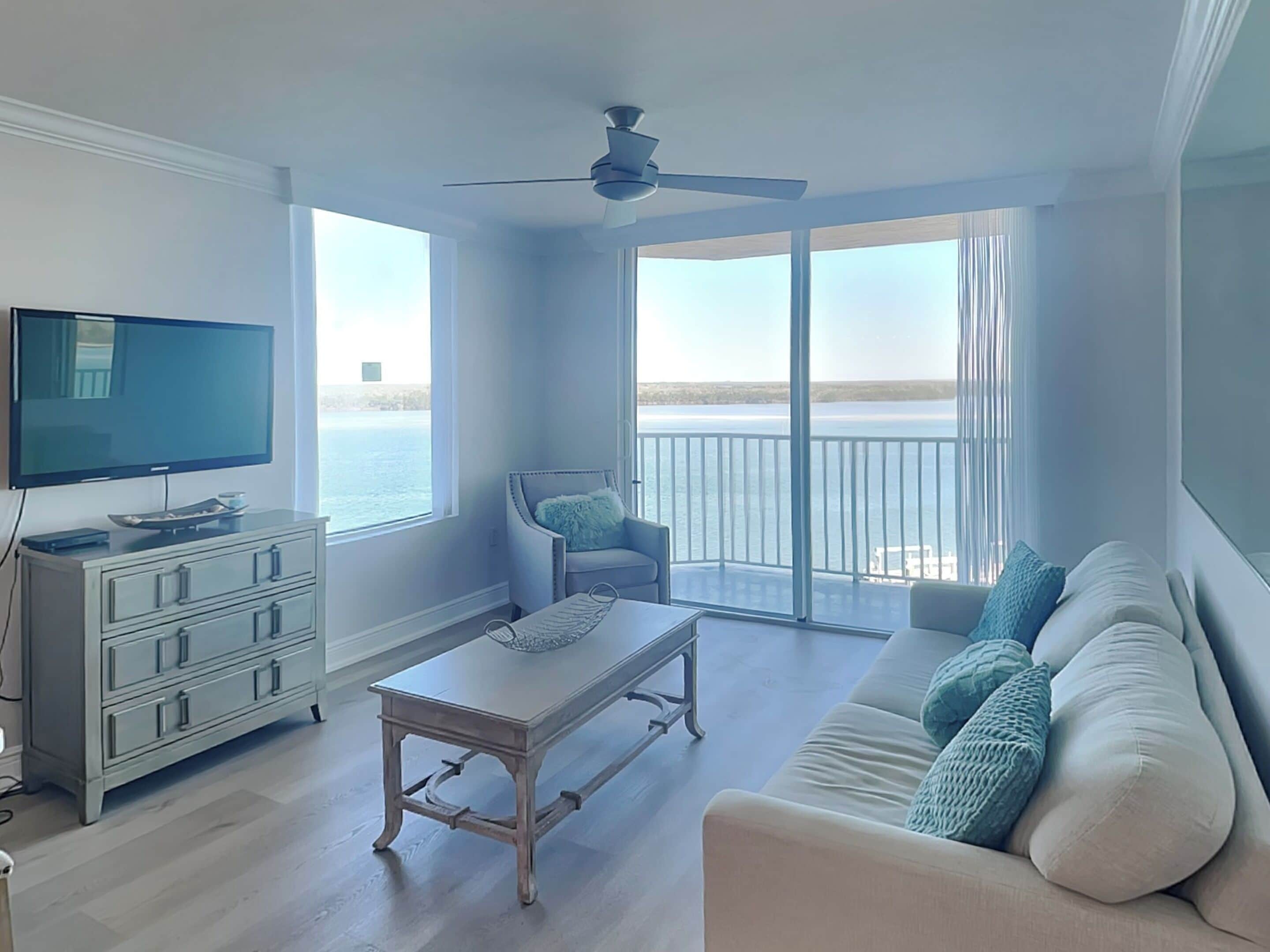 A living room with a television, a sofa, a coffee table, and a chair. There is a balcony with a view of the ocean.