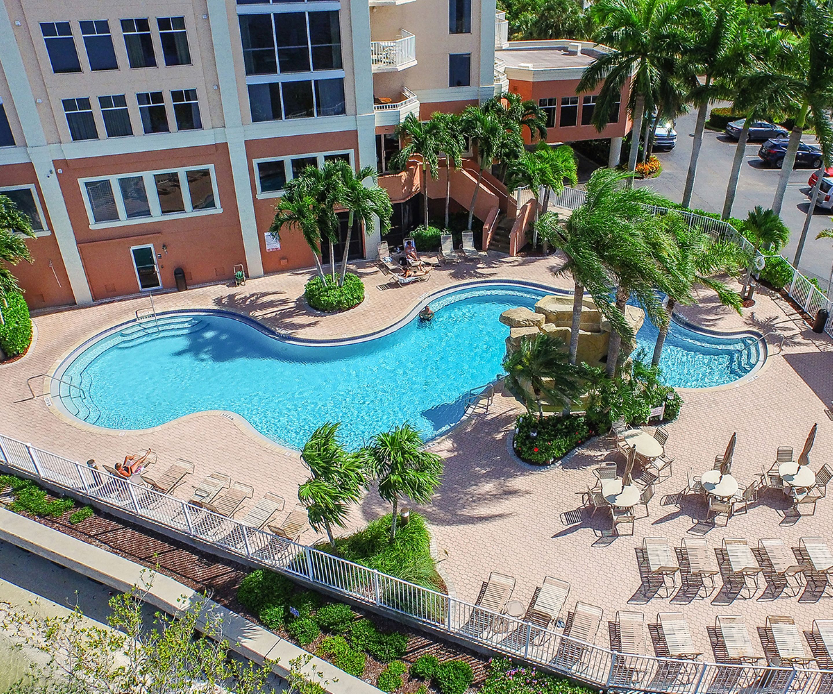 A rooftop swimming pool with lounge chairs and palm trees, surrounded by a multi-story building.