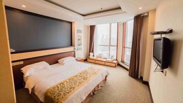 A hotel room with a large bed, a TV, a window with curtains, and a city view. - Nishi Kingmau Hotel (Changzhou)