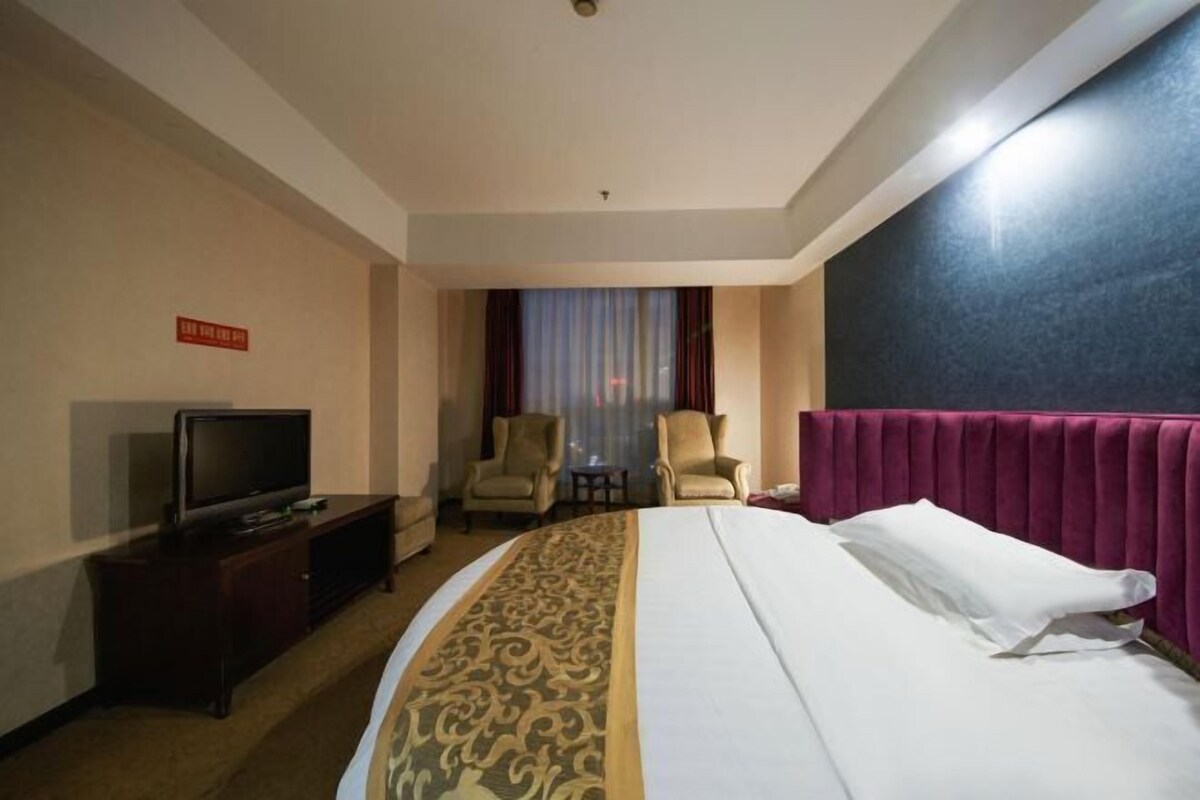 Une chambre d’hôtel avec un lit, un téléviseur posé sur un meuble, deux fauteuils, une petite table et une fenêtre avec des rideaux.
