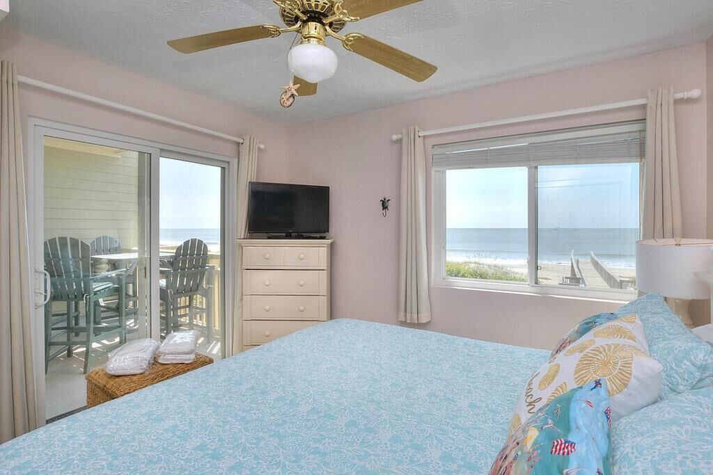 Een slaapkamer met een bed, een televisie, een plafondventilator en uitzicht op het strand.