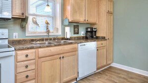 A kitchen with wooden cabinets, a granite countertop, a dishwasher, and a window with blinds. - Charming 2BR duplex with ocean views in Oak Island, perfect for family getaways. (Oak Island)