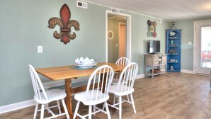 A dining area with a wooden table and white chairs, a decorative wall piece, and a blue shelving unit with decorative items. - Charming 2BR duplex with ocean views in Oak Island, perfect for family getaways. (Oak Island)