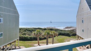 Vista costera con un velero en el agua, rodeado de vegetación de playa y dos edificios con balcón.