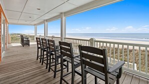 Une terrasse en bois avec des chaises et une table, donnant sur une plage et l’océan.