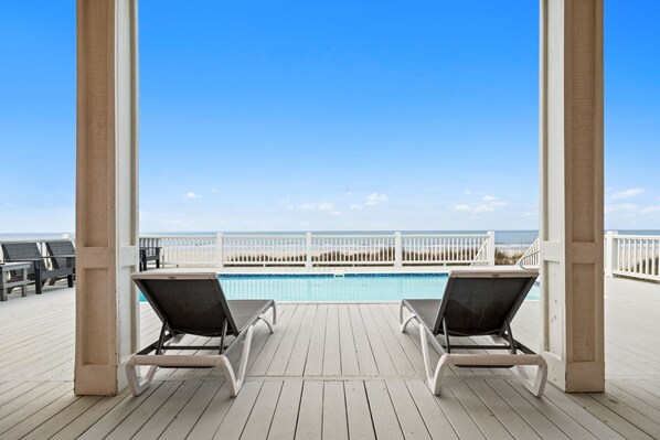A wooden deck with two lounge chairs facing a pool and beach.