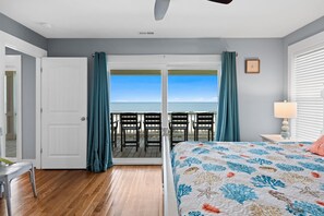 A bedroom with a bed, a nightstand, a lamp, a view of the ocean through a sliding door, and a chair outside.