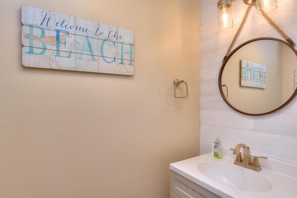 A bathroom with a white sink, a gold faucet, a wooden sign saying "Welcome to the BEACH", and a round mirror with a small framed picture. - A Sandy Feet Retreat - Oceanfront 5-bedroom home in Oak Island (Oak Island)