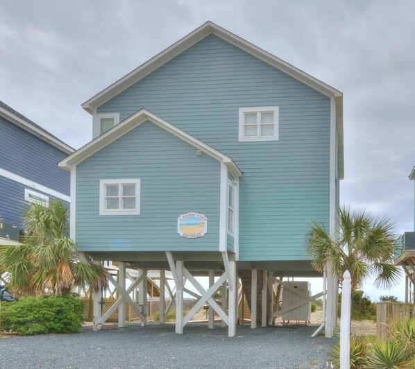 A two-story house with a balcony on stilts, a sign indicating a sandy feet retreat, and palm trees in the background. - A Sandy Feet Retreat - Oceanfront 5-bedroom home in Oak Island (Oak Island)