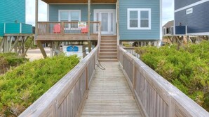A two-story beachfront house with a wooden deck leading to the entrance, a balcony with red chairs, and a sign indicating a rental price.