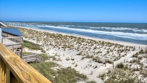 A beach view with a wooden deck and a pier in the background. - Oceans Eleven 03: Stunning 5BR oceanfront home in Oak Island, perfect for families! (Oak Island)