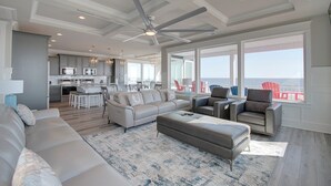 En rummelig opholdsstue med stor sofa, skammel og lænestole, et køkken i baggrunden og en balkon med udsigt.