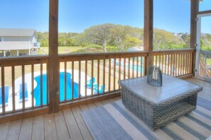 A covered outdoor area with a wooden deck, a wicker table, and a view of a swimming pool and greenery.