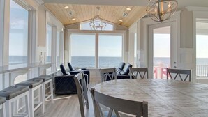 A spacious room with a large wooden table and chairs, a bench with cushions, and large windows offering a view of the ocean.