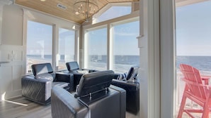 A sunlit room with a view of the ocean, featuring a large window, a ceiling fan, and a seating area with a table.
