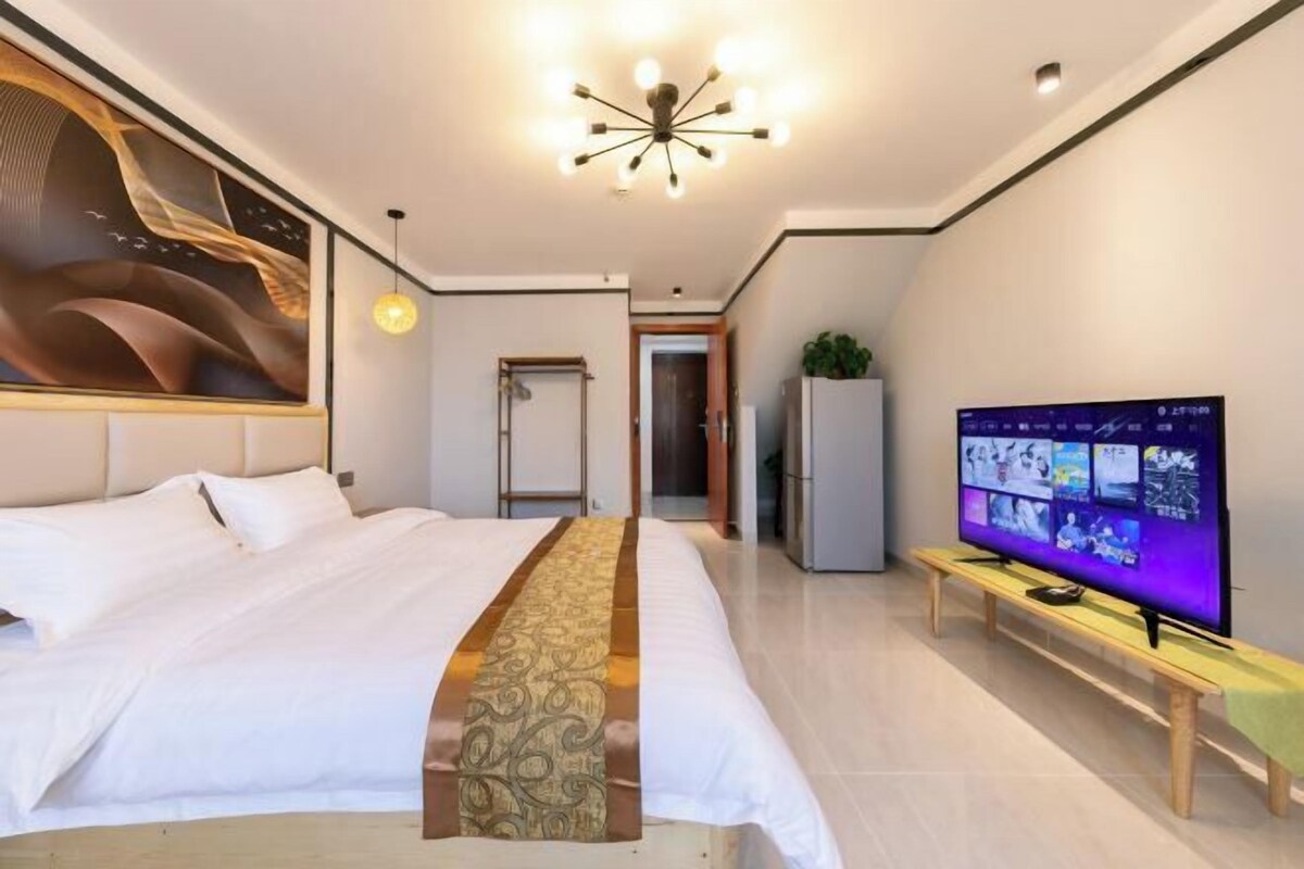 A hotel room with a large bed, a television on a stand, and a hallway leading to a door.