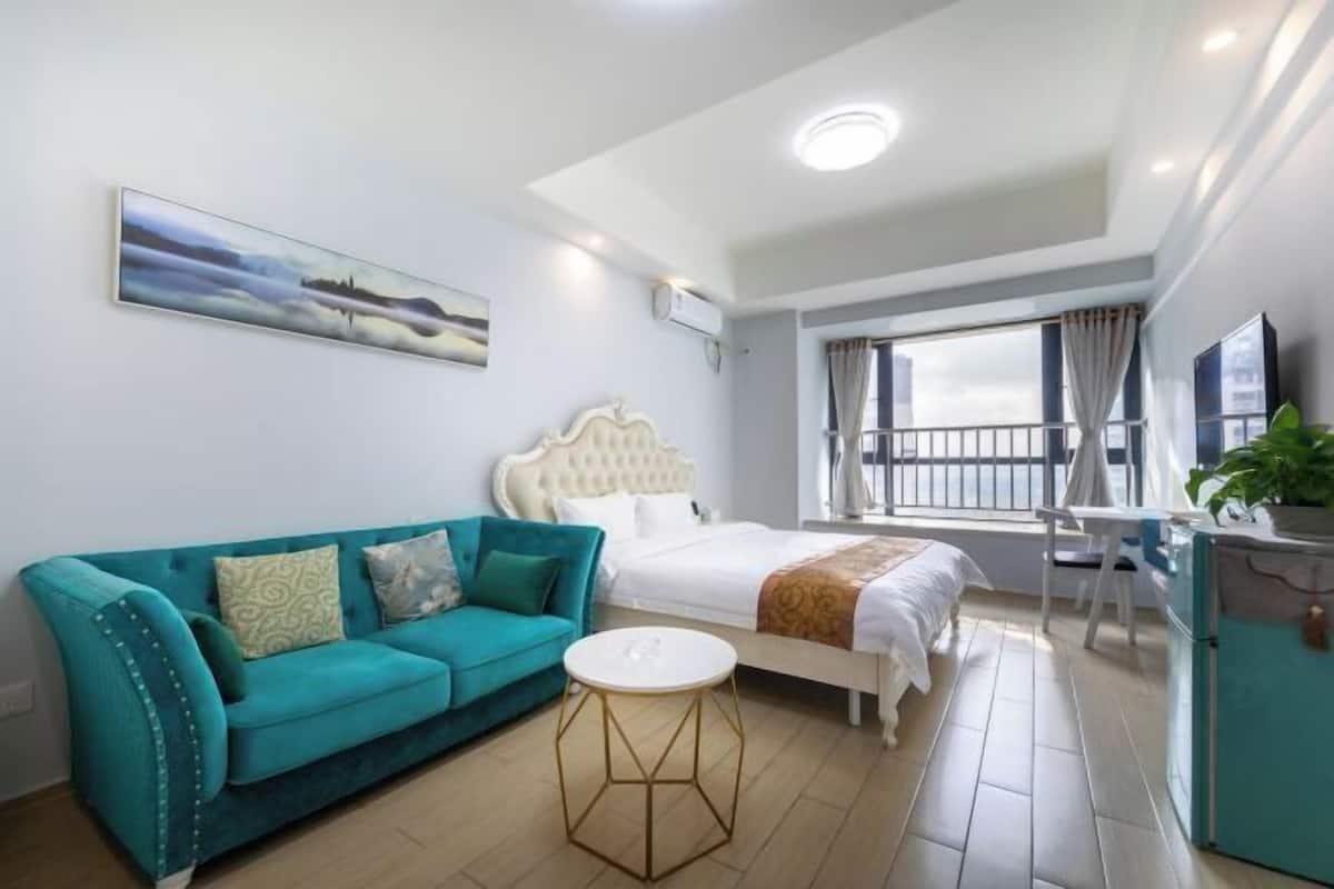 A hotel room with a bed, a sofa, a small round table, a TV, and a balcony with a railing.
