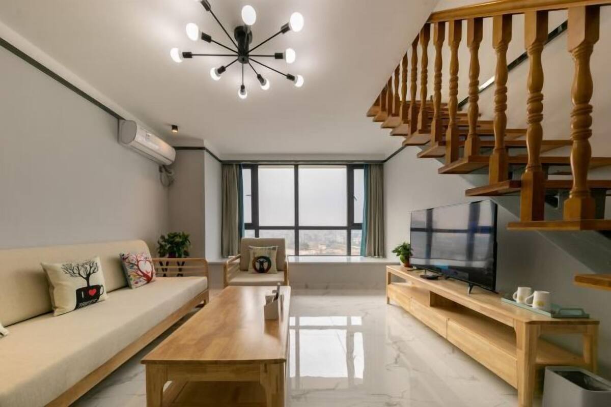 A modern living room with a wooden staircase, a sofa set, a TV, and a large window.