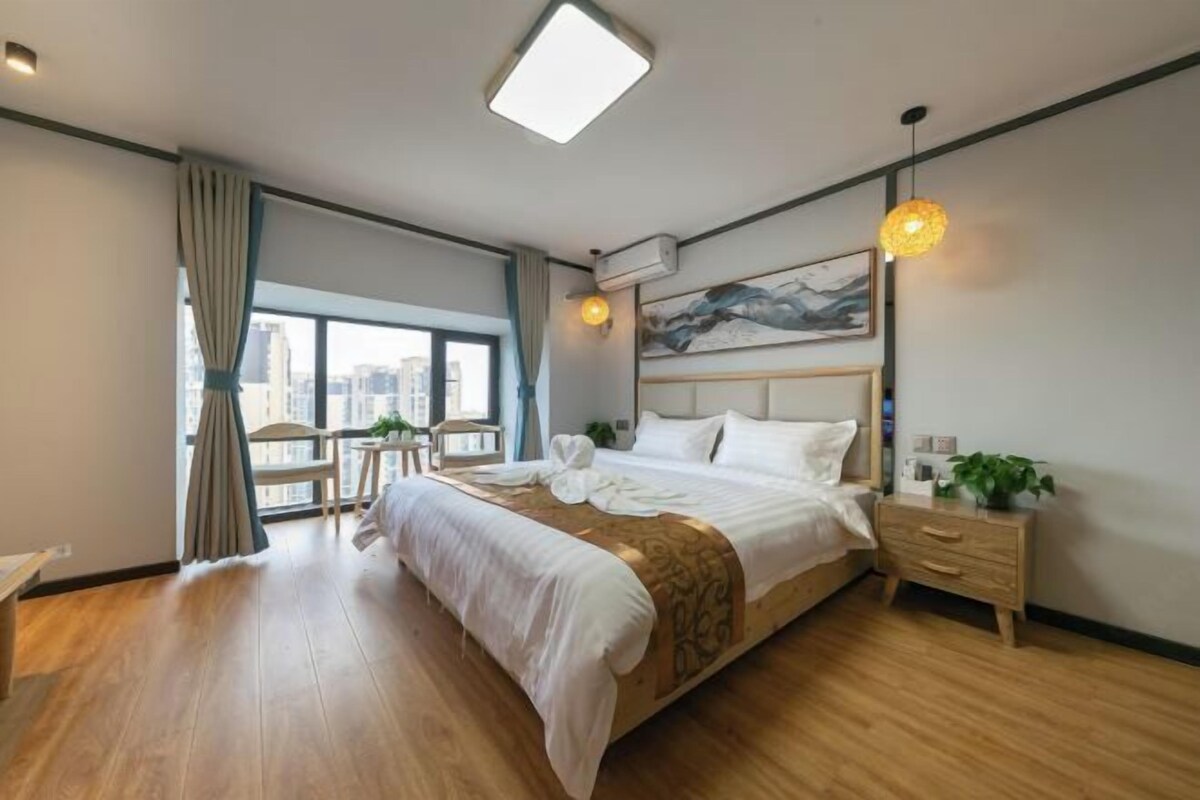 A modern hotel room with a large bed, a wooden floor, a city view, a painting on the wall, and a bedside table with a plant.