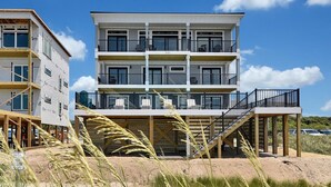 Un edificio a più piani affacciato sulla spiaggia, con balconi, un pontile in legno e la sabbia in primo piano.
